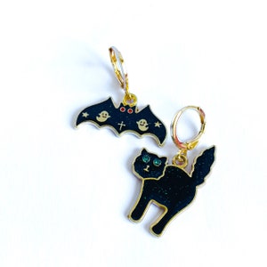 May include: A pair of gold Halloween-themed earrings. One earring features a black bat with a red heart and white ghosts. The other earring features a black cat with green eyes.