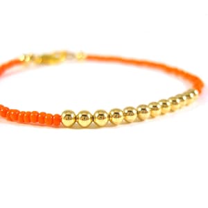 May include: A simple bracelet featuring orange seed beads and gold beads. The bracelet is made with a single strand of beads and has a simple clasp.