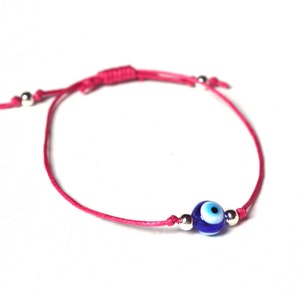 May include: A pink cord bracelet featuring a blue and white evil eye bead. The bracelet has silver-colored beads and an adjustable sliding knot closure. The evil eye bead is a dark blue color with a white and black eye design.