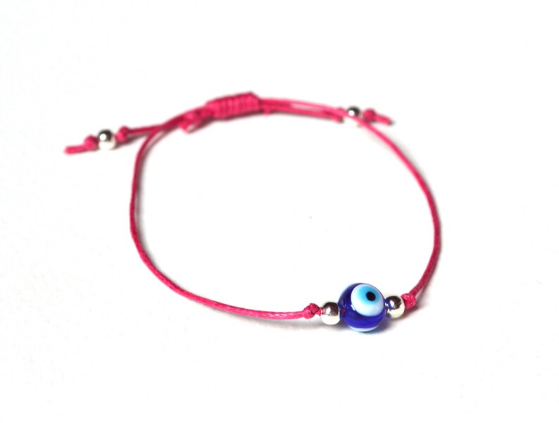 May include: A pink cord bracelet featuring a blue and white evil eye bead. The bracelet has silver-colored beads and an adjustable sliding knot closure. The evil eye bead is a dark blue color with a white and black eye design.