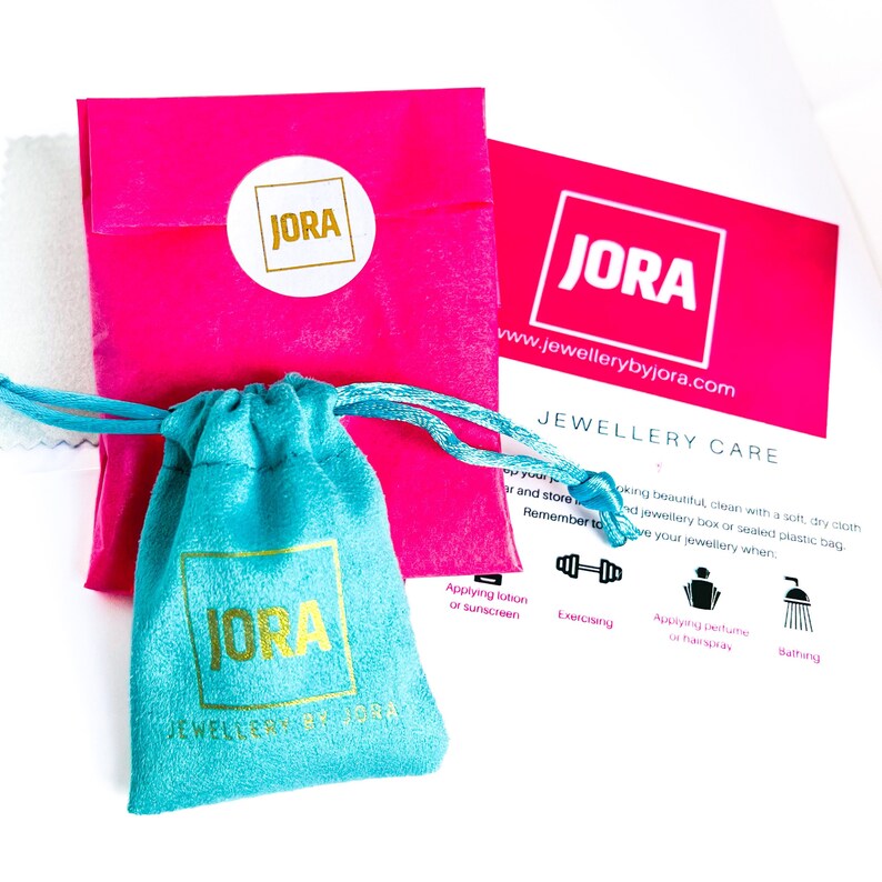May include: A pink velvet pouch with a gold JORA logo, a turquoise velvet pouch with a gold JORA logo, and a white card with the text "JORA" and "www.jewellerybyjora.com" and the heading "JEWELLERY CARE".