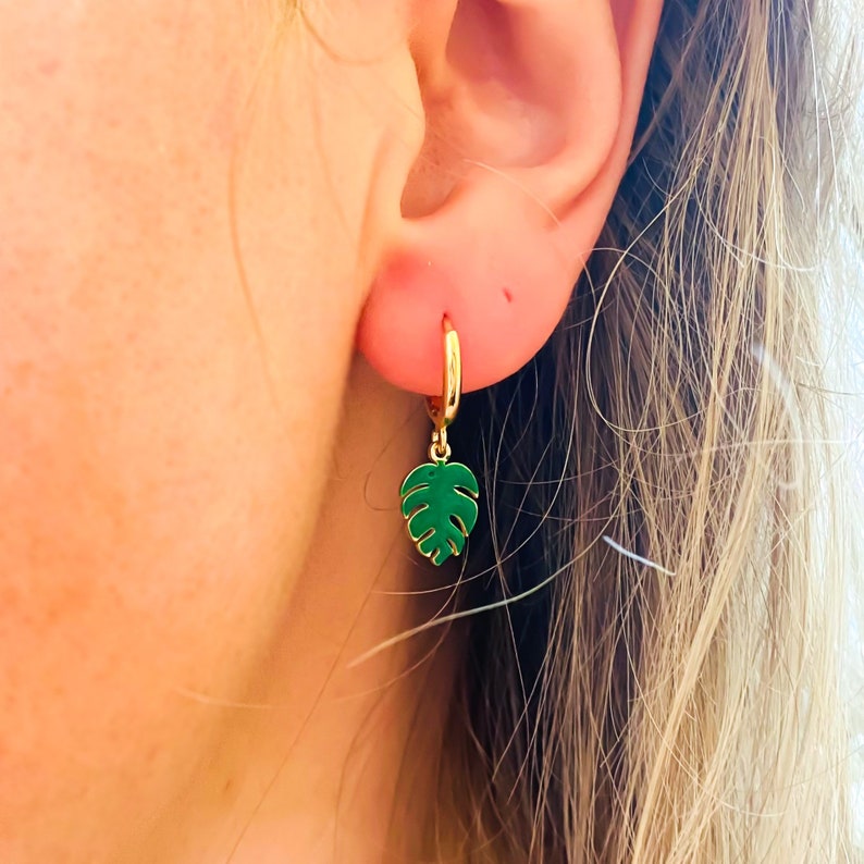 May include: Gold hoop earrings with a green monstera leaf charm hanging from each hoop.