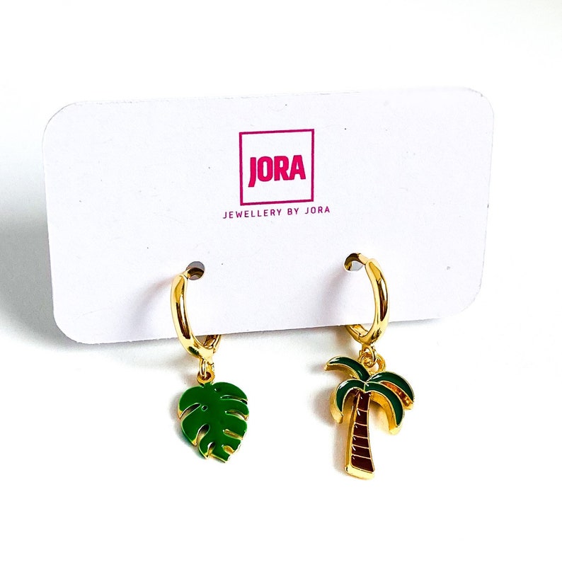 May include: Gold hoop earrings with a green palm tree and a green monstera leaf charm. The earrings are on a white card with the text "JORA JEWELLERY BY JORA".