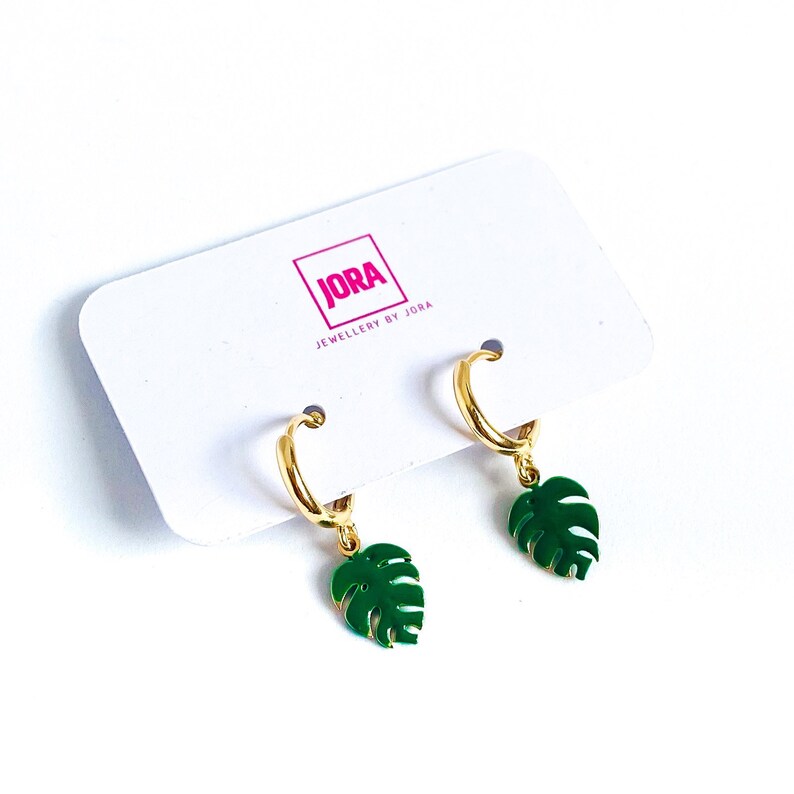 May include: Gold hoop earrings with green monstera leaf charms. The earrings are displayed on a white card with the text "JORA JEWELLERY BY JORA".