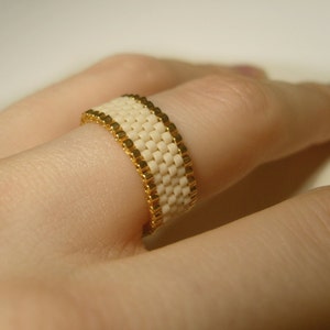 K&ouml;nnte beinhalten: Ein wei&szlig;er Perlenring mit goldenen Akzenten. Der Ring wird an einem Finger getragen.