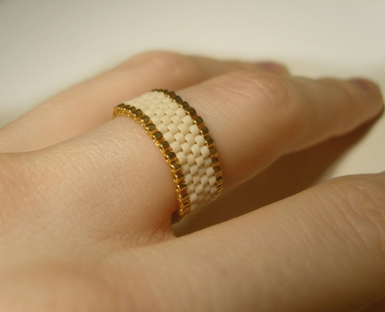 K&ouml;nnte beinhalten: Ein wei&szlig;er Perlenring mit goldenen Akzenten. Der Ring wird an einem Finger getragen.