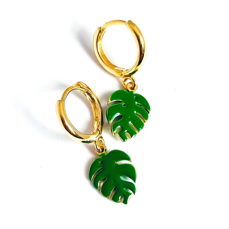 May include: Gold hoop earrings with green monstera leaf charms. The earrings are a simple and stylish design.