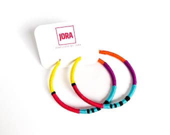 May include: A pair of colorful hoop earrings with a yellow, red, orange, purple, and blue woven design. The earrings are hanging from a white card with the text "JORA JEWELLERY BY JORA".