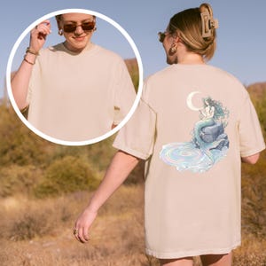 May include: Beige t-shirt featuring a mermaid design on the back. The illustration depicts a mermaid with flowing teal hair, sitting on a rock with a crescent moon above. The shirt has a crew neck and short sleeves.