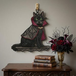 May include: A black cat wall hanging featuring green eyes and fangs, wearing a dark red and black lace cape with a bat-shaped clasp. The cat sits on a decorative black lace shelf. The piece is suspended from a gold-colored wall hook.