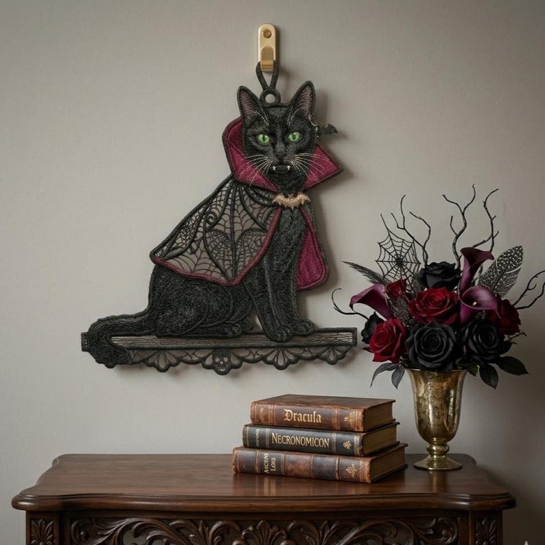 May include: A black cat wall hanging featuring green eyes and fangs, wearing a dark red and black lace cape with a bat-shaped clasp. The cat sits on a decorative black lace shelf. The piece is suspended from a gold-colored wall hook.