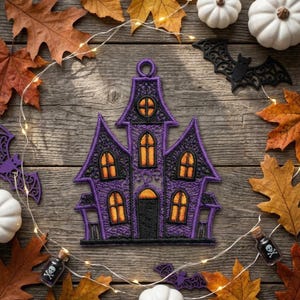 Puede incluir: Un adorno de casa encantada bordado en morado y negro con ventanas de color naranja brillante y un lazo circular para colgar en la parte superior. El dise&ntilde;o presenta detalles de costura intrincados, una puerta central arqueada y varios techos puntiagudos.