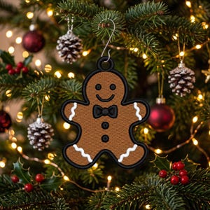 May include: A brown gingerbread man ornament featuring a black embroidered outline, a black bow tie, two black buttons, and white wavy icing details on the arms and legs. The ornament includes a circular loop at the top for hanging.