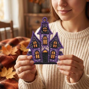 Puede incluir: Un parche bordado en morado y negro con forma de casa encantada, con una torre central, tejados puntiagudos y ventanas iluminadas en naranja. El dise&ntilde;o presenta costuras intrincadas tipo encaje y un peque&ntilde;o lazo circular en la parte superior.