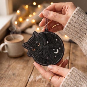 Peut inclure: Un écusson brodé noir de forme circulaire représentant un chat noir enroulé aux yeux ambrés, entouré de petites étoiles blanches brodées et d'un croissant de lune. L'écusson possède une chaîne en métal argenté fixée à l'anneau supérieur.