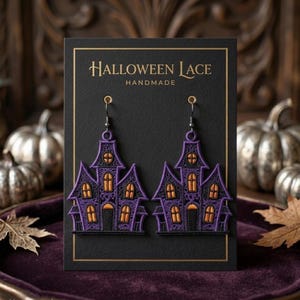 Puede incluir: Un par de pendientes colgantes con forma de casas encantadas, hechos de encaje morado y negro con ventanas naranjas. Los pendientes se presentan en una tarjeta negra con el texto HALLOWEEN LACE HANDMADE en letras doradas.