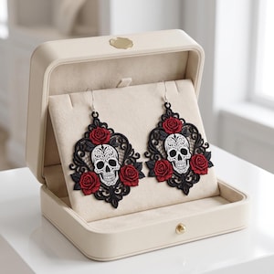 May include: A pair of dangling earrings featuring intricate black lace filigree, white sugar skull motifs, and vibrant red roses. The earrings are displayed inside a cream-coloured jewellery box with a soft velvet interior lining and a gold clasp.