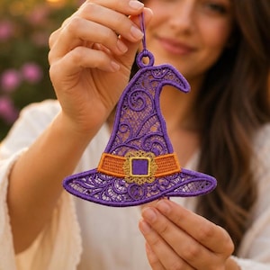 May include: A purple lace witch hat ornament featuring intricate swirling patterns, an orange band, and a gold square buckle. The decorative piece includes a small loop at the top for hanging and is crafted with detailed embroidery work throughout.
