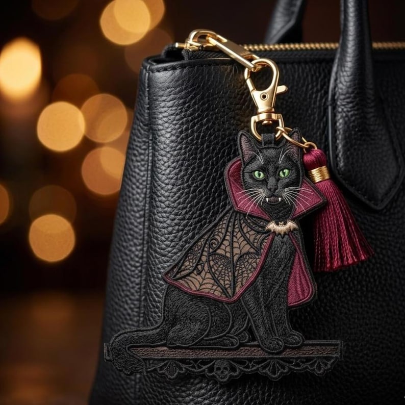 May include: A black leather handbag features a decorative embroidered keychain of a black cat dressed as a vampire. The cat wears a maroon-lined cape with a bat-wing pattern and a small bat charm, accompanied by a matching maroon tassel.