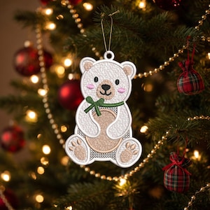 May include: A white embroidered bear ornament with a tan belly, ears, and paw pads, featuring pink cheeks, a black nose, and a green ribbon tied around its neck. The bear is suspended by a thin gold string and has a white mesh-like border.