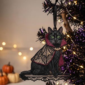 May include: A black cat ornament with green eyes and white fangs, wearing a maroon and tan cape with a spiderweb pattern and a small bat brooch. The cat sits on a decorative shelf featuring a skull motif, suspended by a black ribbon.