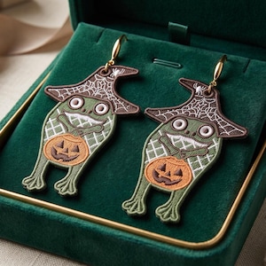 May include: A pair of embroidered earrings featuring green frogs wearing brown witch hats with white spiderweb patterns. Each frog holds an orange jack-o'-lantern pumpkin. The earrings are attached to gold-toned metal hooks for pierced ears.