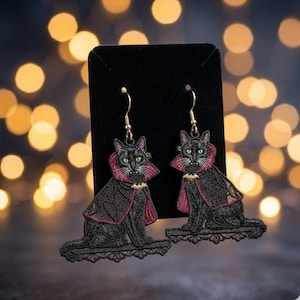 May include: A pair of dangling earrings featuring embroidered black cats wearing red-lined capes with gold bat-shaped clasps. The cats have green eyes and visible fangs, and each sits on a black lace-like base attached to gold-toned hooks.
