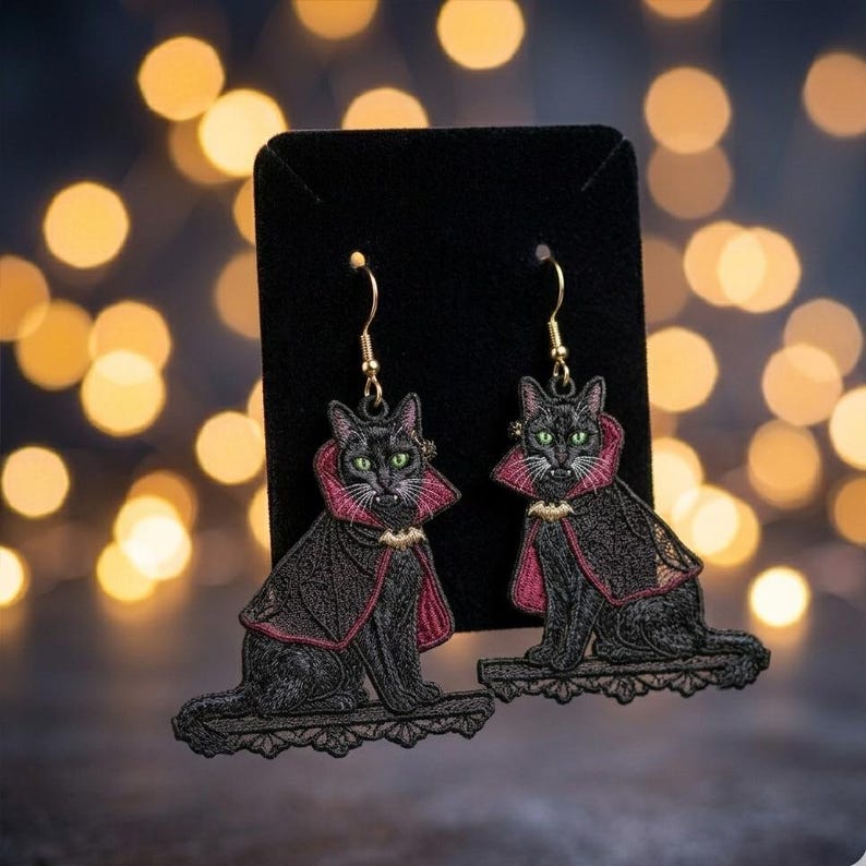 May include: A pair of dangling earrings featuring embroidered black cats wearing red-lined capes with gold bat-shaped clasps. The cats have green eyes and visible fangs, and each sits on a black lace-like base attached to gold-toned hooks.