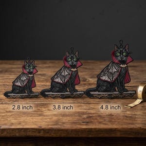 May include: Three black cat embroidered ornaments with green eyes and fangs, wearing red and white spiderweb capes. The ornaments are labeled 2.8 inch, 3.8 inch, and 4.8 inch, with a measuring tape visible next to the largest cat.