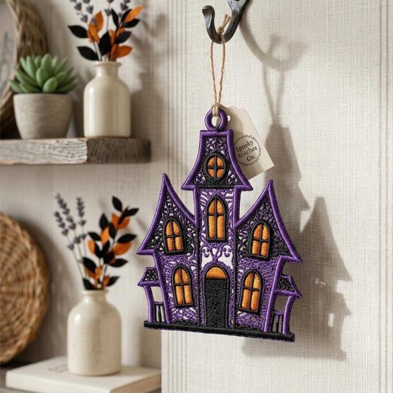 Puede incluir: Un adorno bordado de casa encantada en color morado y negro con ventanas naranjas y una puerta central. El dise&ntilde;o incluye costuras intrincadas tipo encaje y est&aacute; suspendido por un cordel fino con una etiqueta que dice Spooky Stitchee Co.