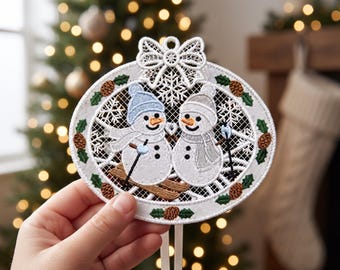 Puede incluir: Un adorno navideño de encaje blanco con dos muñecos de nieve bordados sobre esquís. Los muñecos llevan gorros y bufandas azules y grises. El marco circular está decorado con hojas de acebo verdes y piñas marrones, con un lazo de encaje.