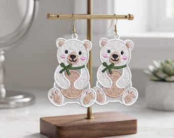 Puede incluir: Un par de pendientes de oso bordados en blanco con barriga color canela, mejillas rosadas y una pajarita verde oscuro. Los osos tienen ojos y nariz negros, con costuras detalladas. Los pendientes cuelgan de ganchos de metal dorado.