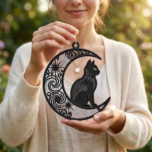 Könnte beinhalten: Ein schwarzes, besticktes Ornament in Form eines Halbmonds mit einer sitzenden schwarzen Katze mit grünen Augen. Der Mond ist mit komplizierten Wirbelmustern und silbernen Sternmotiven verziert. Das Stück ist kreisförmig und hat eine kleine Schlaufe zum Aufhängen, die detaillierte Handarbeit und kontrastierende silberne Fadenakzente zeigt.