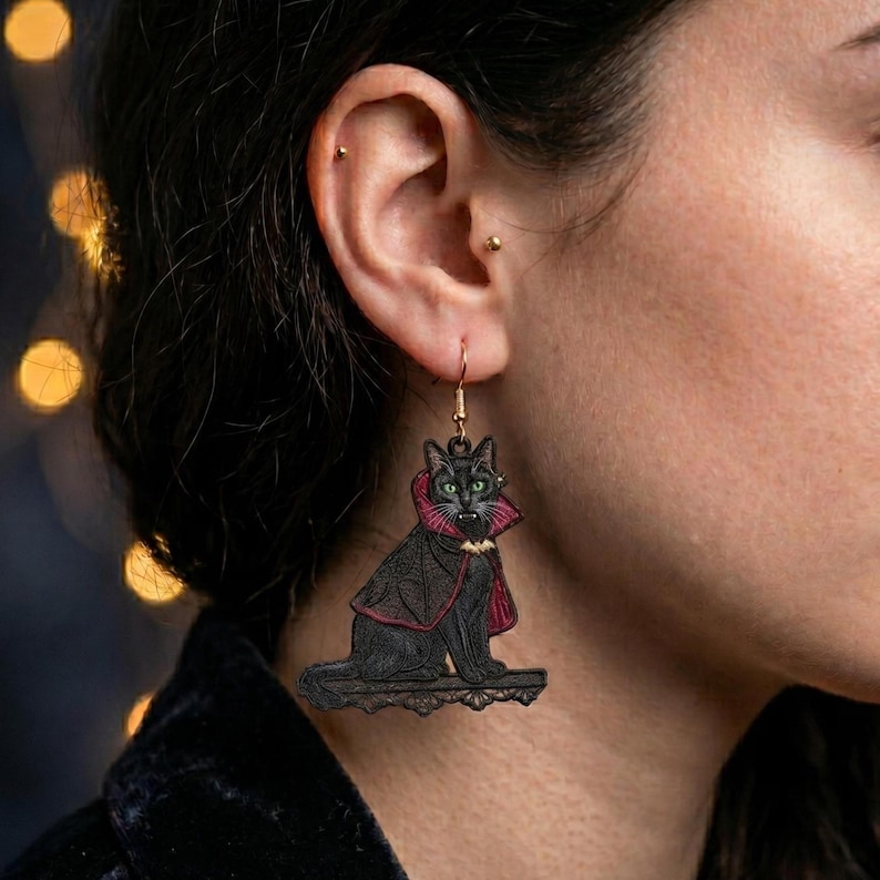 May include: A dangling earring featuring a black cat illustration wearing a red-lined black vampire cape with a small bat emblem at the collar. The cat has green eyes and is sitting on a decorative ledge, suspended from a gold-toned hook.