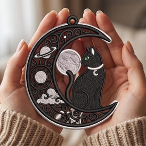 May include: A circular black and white embroidered ornament featuring a crescent moon frame with intricate scrollwork. Inside the frame is a black cat with green eyes, a white collar, a planet with rings, a sun, and a cloud motif.