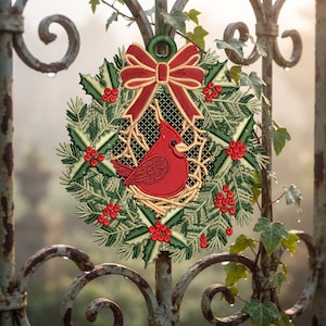 May include: A circular embroidered ornament featuring a red cardinal bird centered within a wreath of green holly leaves and red berries. A red bow with gold trim sits at the top, and the design is set against a black mesh background.