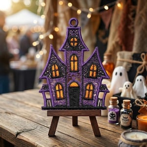 May include: A purple and black embroidered haunted house ornament featuring orange glowing windows and a central door, displayed on a small wooden easel. The intricate lace-like stitching forms the structure of the house with a circular loop at the top.