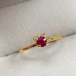 Può includere: Un delicato anello d'oro con una gemma rossa rotonda e profonda. L'anello ha un piccolo elemento decorativo con minuscole pietre trasparenti. L'anello è presentato su uno sfondo bianco.
