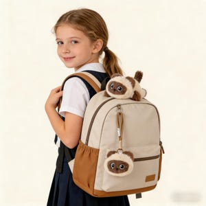 May include: A beige backpack with brown detailing, adorned with a matching plush cat toy and keyring charm. The backpack includes a front pocket and a top zip. The plush cat is brown and beige. Ideal for school or general use.