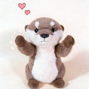 Puede incluir: Un peluche de nutria con pelaje marr&oacute;n y blanco, brazos extendidos y nariz negra. Dos corazones rosas flotan sobre la nutria. El juguete est&aacute; dise&ntilde;ado para parecer amigable e invitador.