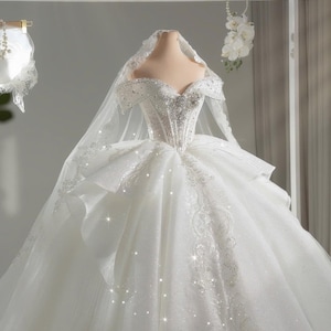 Op de afbeelding: Een witte trouwjurk met een sweetheart-halslijn en een volle, gelaagde rok. Het lijfje is versierd met sprankelende kristallen en gedetailleerd kralenwerk. Een doorschijnende sluier bedekt de schouders. De jurk wordt op een mannequin gepresenteerd.