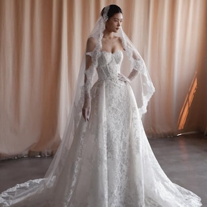 Puede incluir: Vestido de novia de encaje blanco con escote corazón y cola larga. El vestido está adornado con intrincados patrones de encaje floral. Un velo y guantes a juego completan el look nupcial.
