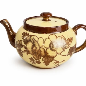 May include: A round ceramic teapot featuring a cream-colored body with a dark brown floral pattern. The teapot includes a matching lid with a small knob, a curved spout, and a handle, all finished in a glossy dark brown glaze that matches the floral design.