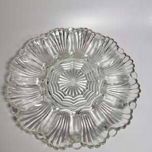 May include: A clear glass serving platter featuring a central circular section with a sunburst pattern, surrounded by eight distinct shell-shaped compartments. The outer edge has a scalloped rim design with small circular indentations between each shell.