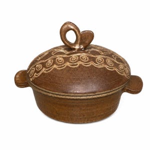 May include: A brown ceramic lidded casserole dish with two side handles. The lid features a decorative embossed swirl pattern and a central loop-shaped handle. The surface has a speckled texture and a glossy finish throughout the entire piece.
