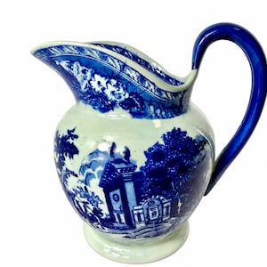 May include: A ceramic pitcher featuring a white base with blue transferware patterns depicting a landscape with buildings and trees. The pitcher has a curved handle, a wide spout, and a decorative floral border along the upper rim.