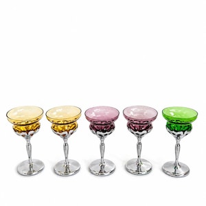 May include: Five colorful stemmed glasses with a silver base and stem. The glasses have a rounded bowl in shades of yellow, pink, and green. The glasses are arranged in a row against a white background.