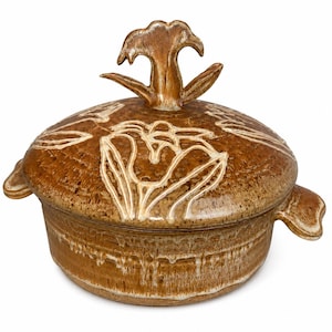 May include: A round, brown ceramic lidded casserole dish with a speckled glaze finish. The lid features a sculpted flower handle and an incised floral pattern. Two small handles extend from the sides of the base, which has a drip glaze effect.