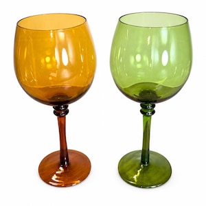 Puede incluir: Dos copas de vino con pie. Una es de color ámbar, la otra verde. Ambas tienen un cuenco redondeado y un tallo largo. La copa ámbar tiene un tallo y una base marrones, mientras que la copa verde tiene un tallo y una base verdes. Sobre fondo blanco.