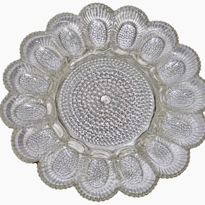 May include: A clear glass egg serving platter featuring a circular center with a textured hobnail pattern, surrounded by twelve individual oval-shaped indentations with ribbed edges designed to hold eggs in a radial arrangement.